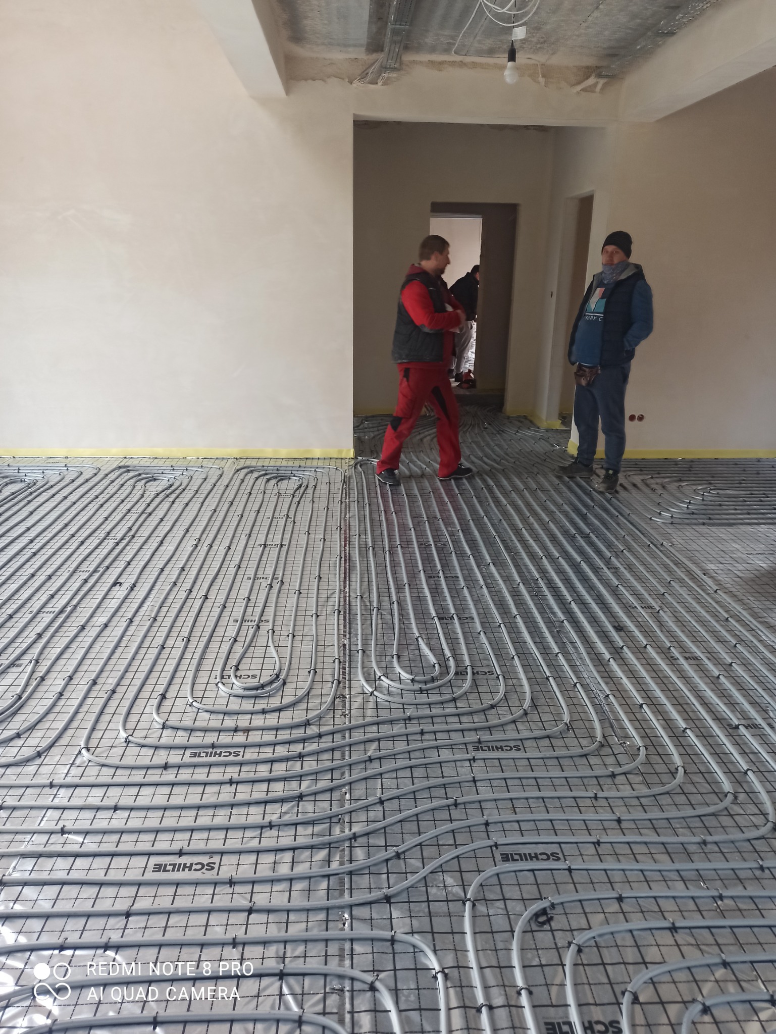 Szare rury ogrzewania podłogowego ułożone na folii z nadrukiem Schilte, widoczni dwaj instalatorzy w tle, jeden w czerwonym kombinezonie, drugi w niebieskiej kurtce i czapce.