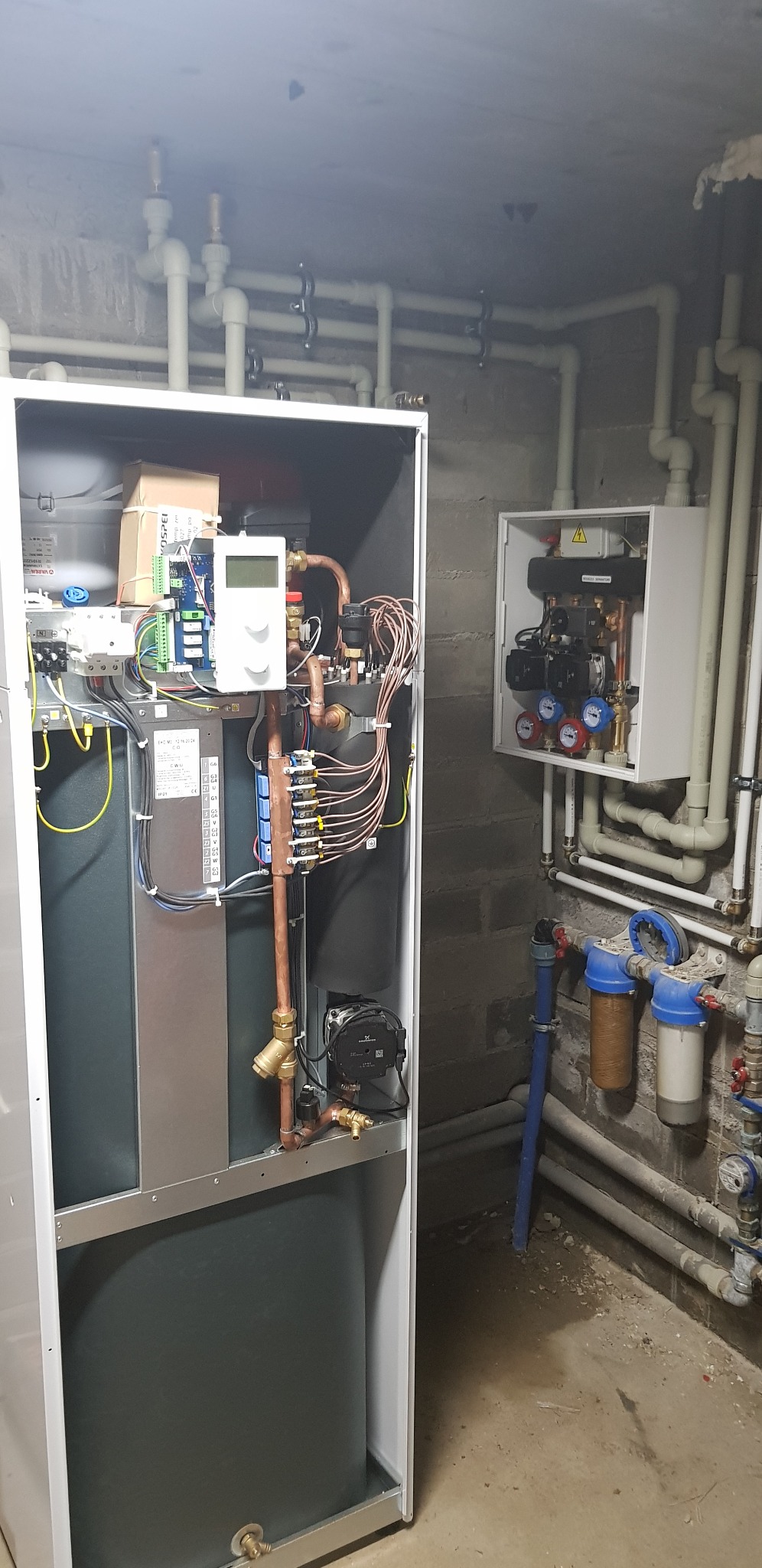 Wnętrze kotłowni z nowoczesnym piecem kondensacyjnym w białej obudowie, widoczne liczne rury miedziane i plastikowe, sterownik elektroniczny oraz dodatkowy panel sterujący na ścianie.