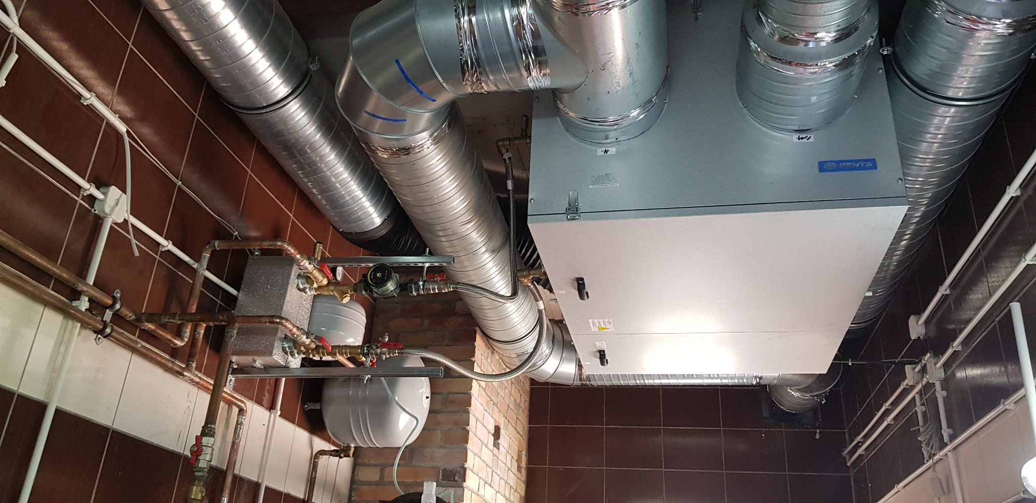 Centrala wentylacyjna z podłączonymi przewodami wentylacyjnymi i instalacją hydrauliczną na tle brązowych płytek.
