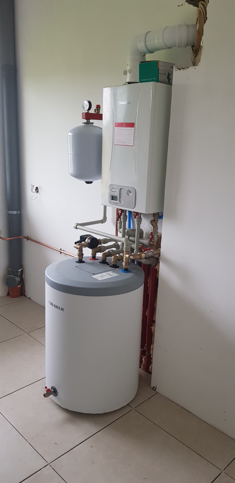 Biały bojler z szarym wykończeniem, podłączony do pieca gazowego Vaillant, z widocznymi rurami miedzianymi i białymi plastikowymi, zamontowany na kafelkach w pomieszczeniu z surowymi ścianami...