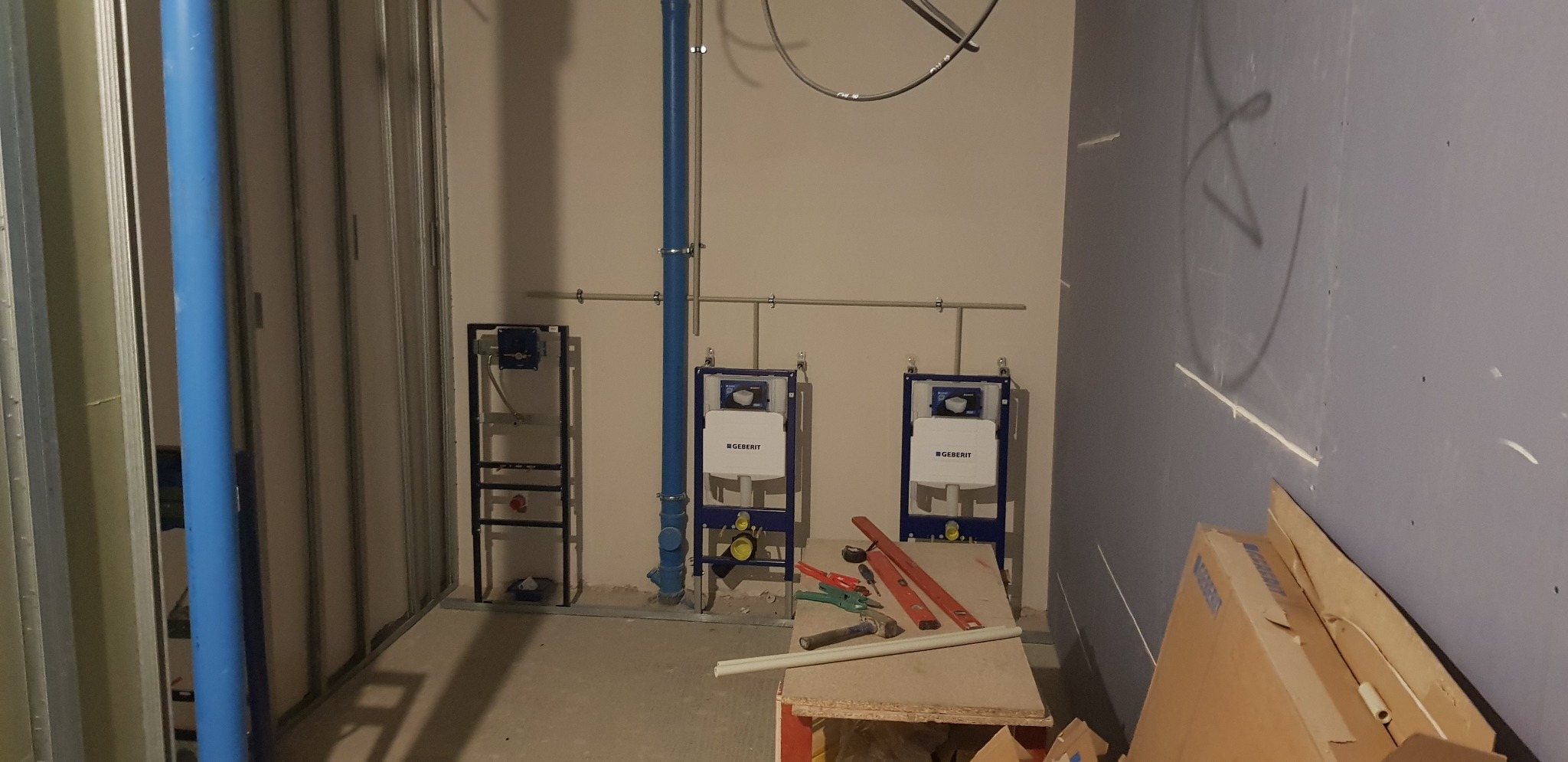 Stelaże podtynkowe Geberit zamontowane na ścianie, widoczne niebieskie rury instalacyjne, poziomice i narzędzia na prowizorycznym stole roboczym, surowy stan wykończenia wnętrza.
