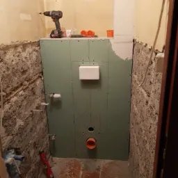 Surowa łazienka w trakcie prac: ściany z odsłoniętą cegłą, częściowo zakryte zieloną płytą kartonowo-gipsową z widoczną instalacją hydrauliczną, wiertarka na płycie, poziomica na podłodze.