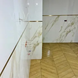 Wykończona łazienka z marmurowymi płytkami na ścianach z cienkim złotym paskiem i drewnopodobną podłogą ułożoną w jodełkę, widoczne wystające kable i rury przed montażem armatury.