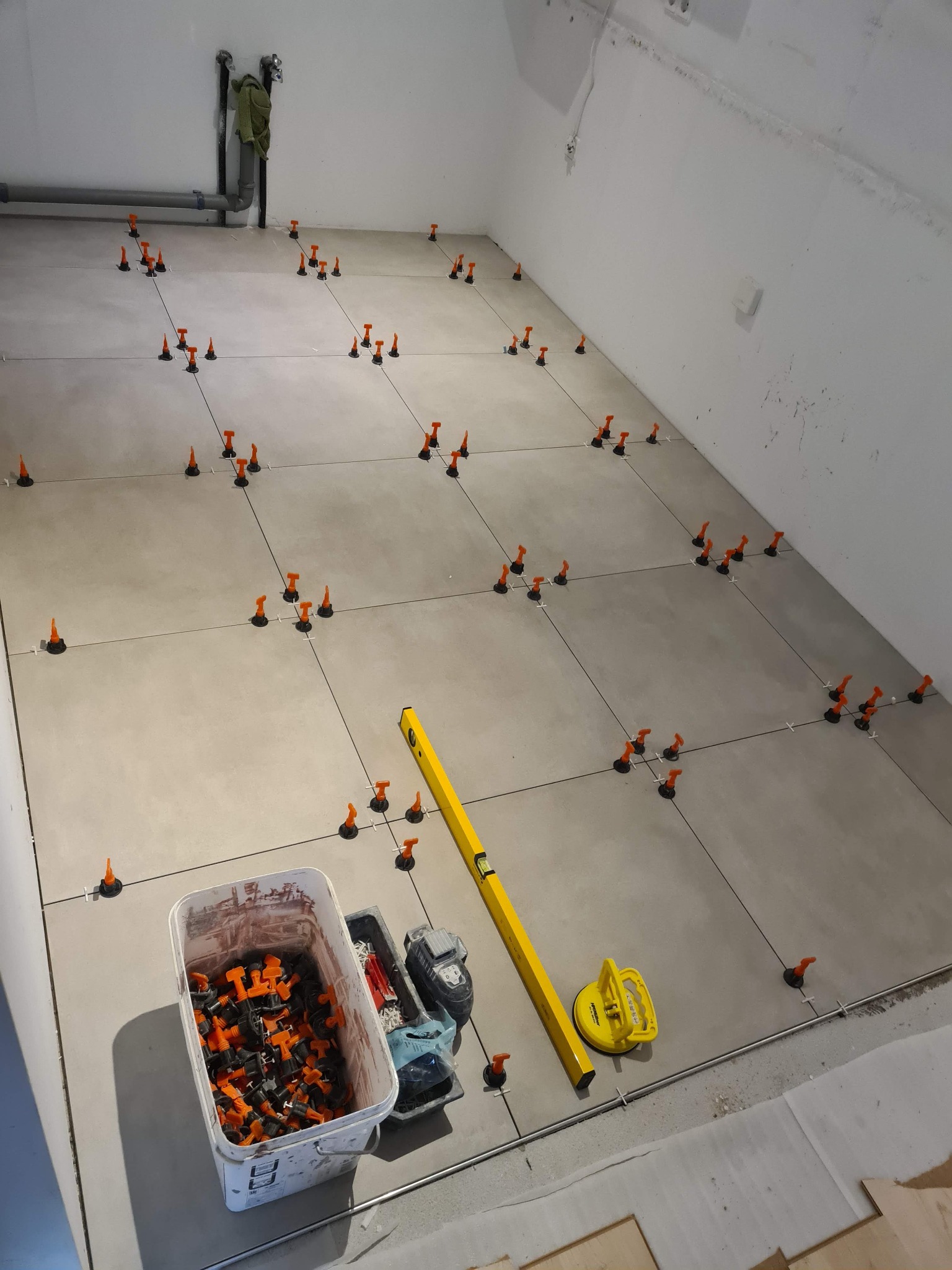 Układanie płytek podłogowych z użyciem poziomicy, przyssawki do płytek i systemu poziomowania płytek z pomarańczowymi klipsami, widoczne rury instalacyjne przy suficie.
