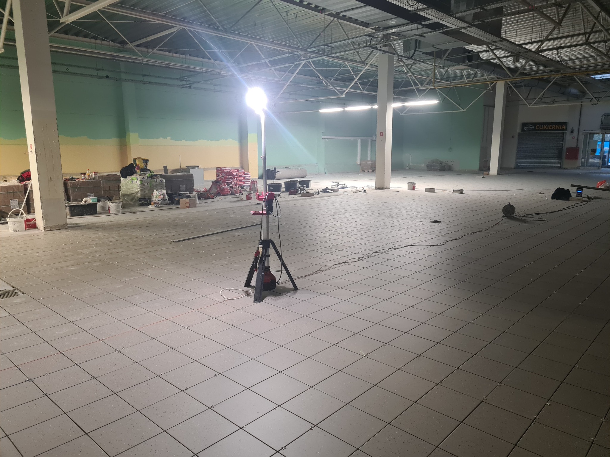 Układanie szarej terakoty w dużej hali przemysłowej oświetlonej lampą budowlaną, widoczne materiały budowlane i narzędzia w tle.