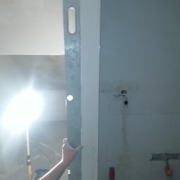 Daniel Czerwiński - Metalowa łata w dłoni osoby z tatuażem, sprawdzająca pion ściany w trakcie prac wykończeniowych, widoczne kable elektryczne i lampa budowlana w tle.