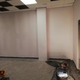 Daniel Czerwiński - Świeżo pomalowane na biało ściany biura z widoczną taśmą malarską przy suficie, podłodze i kolumnie, z narzędziami malarskimi zabezpieczonymi folią na podłodze oraz żółtym kaskiem w rogu...