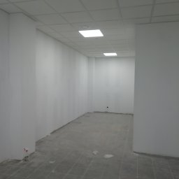 Daniel Czerwiński - Pomalowane na biało wnętrze biura z kasetonowym sufitem i szarą podłogą w trakcie wykańczania, widoczna butelka ze spryskiwaczem przy ścianie.