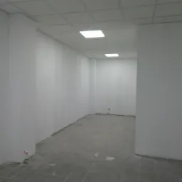 Pomalowane na biało wnętrze biura z kasetonowym sufitem i szarą podłogą w trakcie wykańczania, widoczna butelka ze spryskiwaczem przy ścianie.