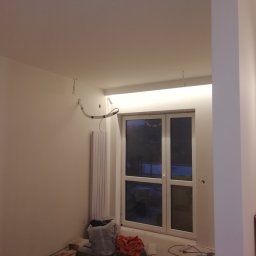 Daniel Czerwiński - Wnętrze pokoju w trakcie prac wykończeniowych, widoczne świeżo pomalowane ściany, grzejnik, okno i przewody elektryczne wystające ze ściany i sufitu.