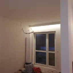 Wnętrze pokoju w trakcie prac wykończeniowych, widoczne świeżo pomalowane ściany, grzejnik, okno i przewody elektryczne wystające ze ściany i sufitu.