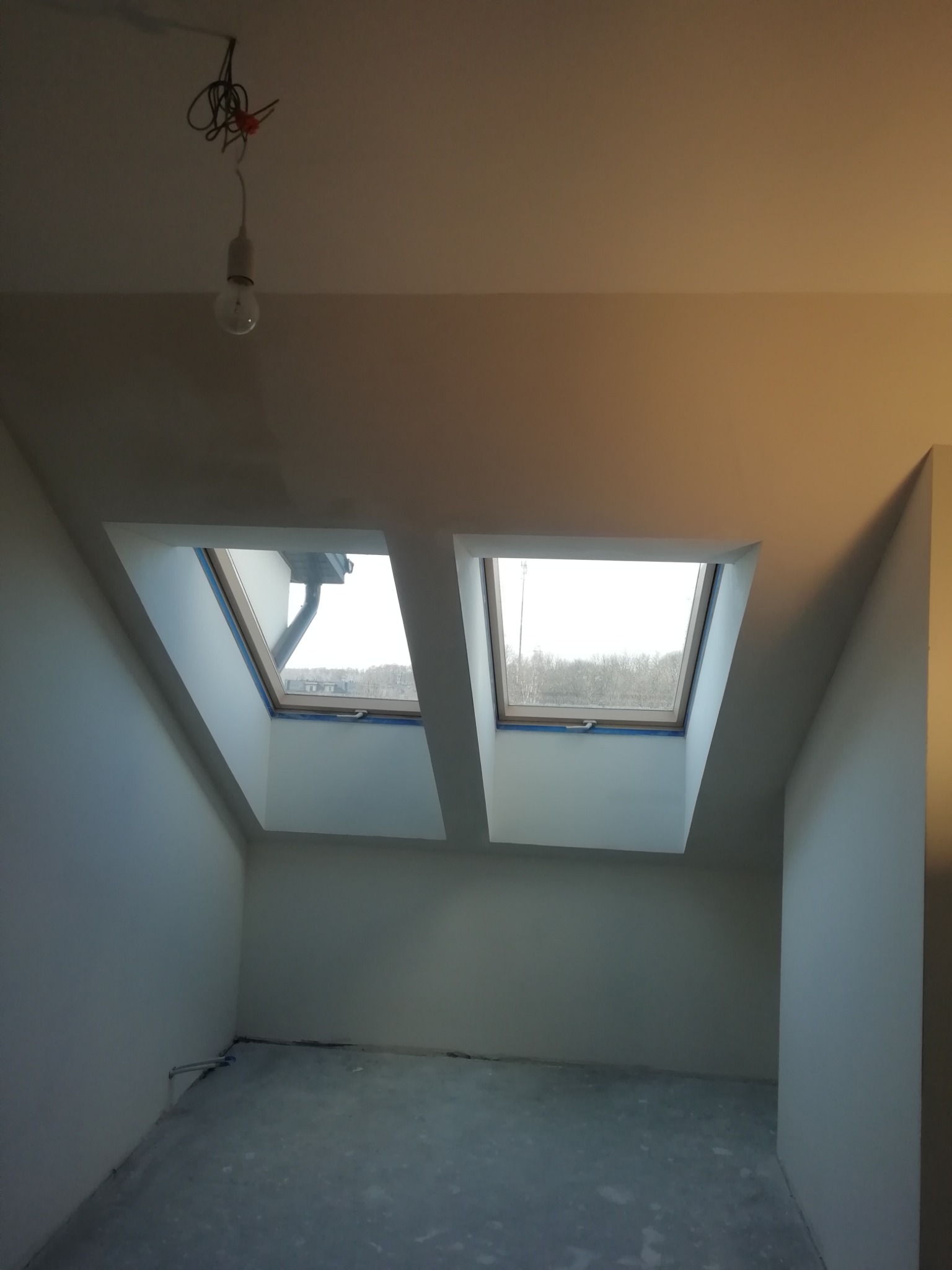 Pokój na poddaszu z dwoma oknami dachowymi, zabezpieczonymi niebieską taśmą malarską, surowy beton na podłodze, widoczna żarówka na kablu.