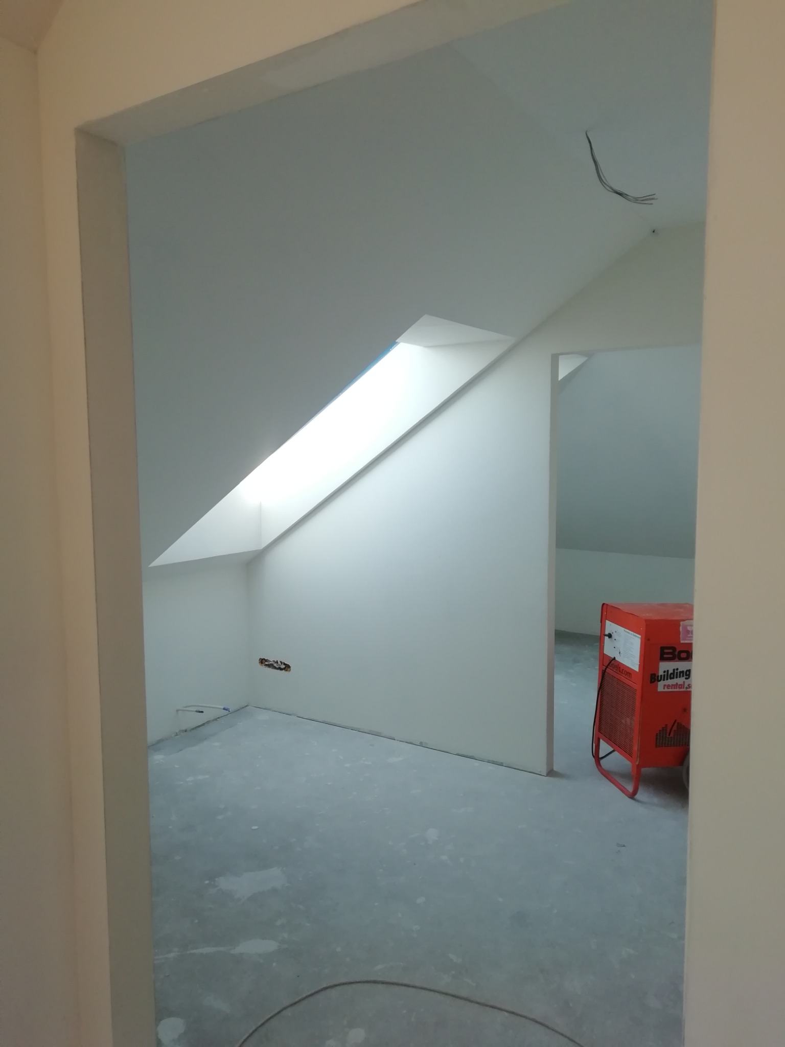 Pokój na poddaszu po świeżym malowaniu na biało, widoczne okno dachowe, wystające kable ze ściany i nagrzewnica budowlana.