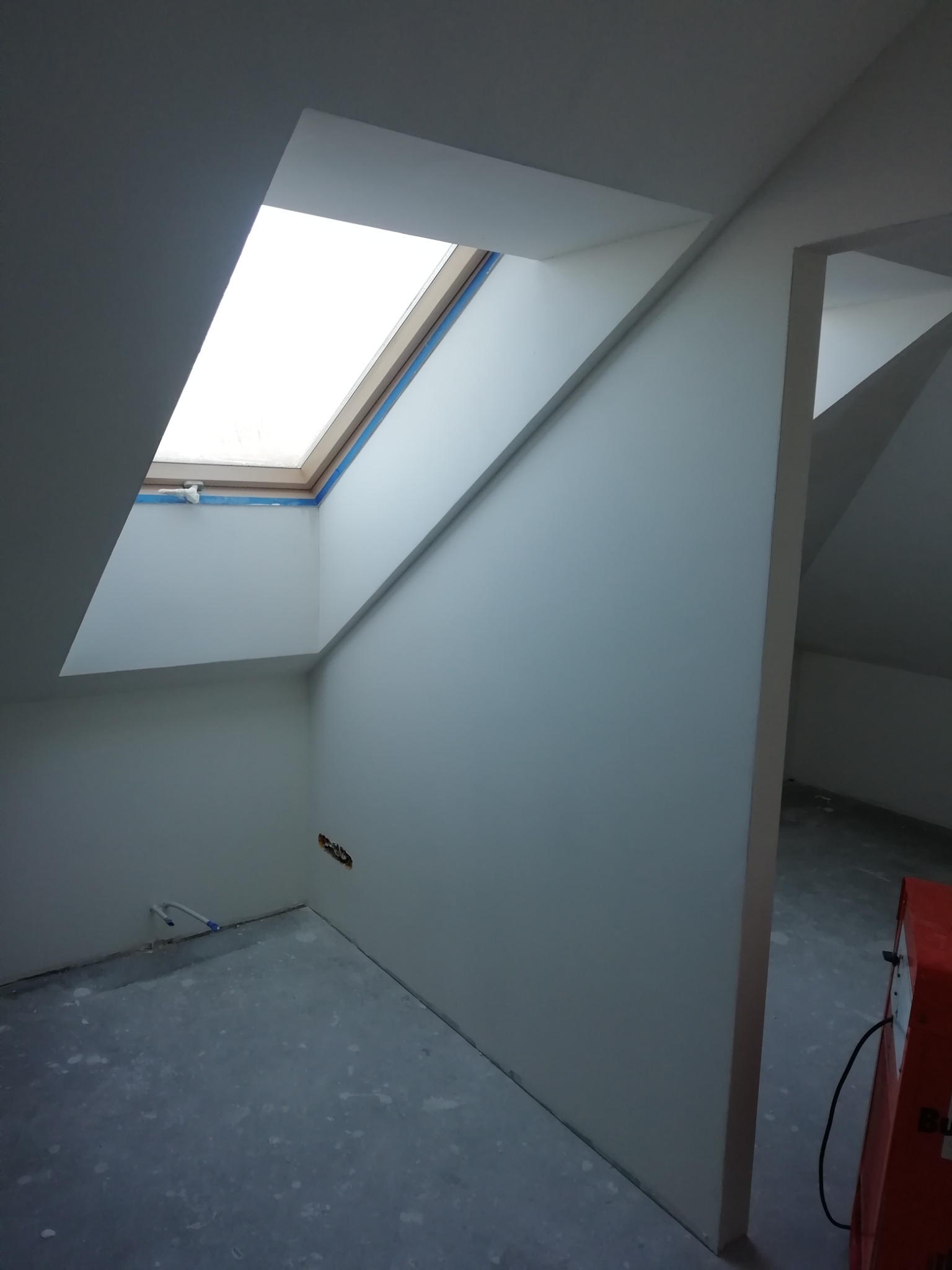 Poddasze po wygładzeniu ścian, widoczne okno dachowe zabezpieczone taśmą malarską, instalacja elektryczna i rury w podłodze, w tle suszarka przemysłowa.