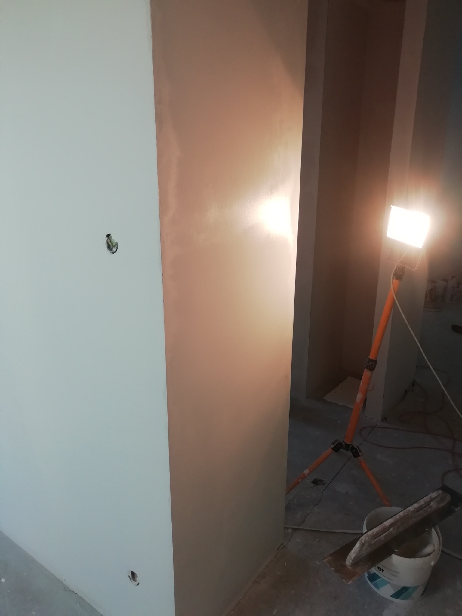 Świeżo nałożona gładź na ścianie, oświetlona lampą budowlaną na trójnogu, wiadro z zaprawą i szpachelka w rogu pomieszczenia, widoczne otwory na gniazdka elektryczne w ścianie.