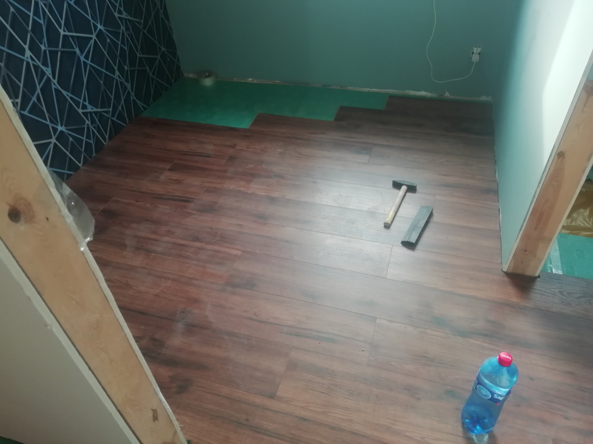 Proces układania paneli podłogowych w Łodzi: widoczny fragment podkładu, ułożone panele imitujące drewno, młotek, dobijak i butelka wody.