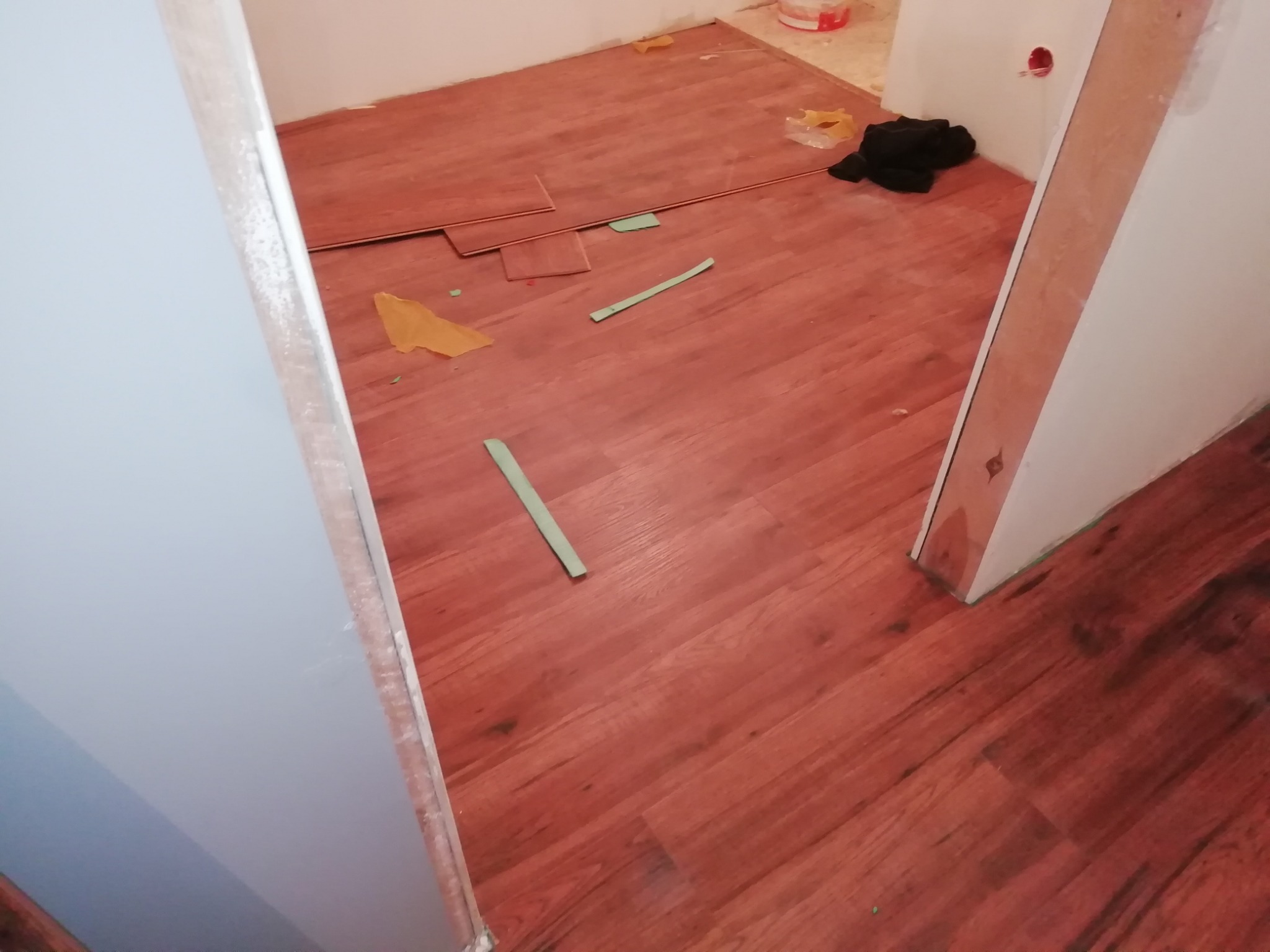 Proces układania paneli podłogowych w odcieniach czerwieni, widoczne fragmenty paneli, listwy przypodłogowe, resztki folii i narzędzia, ujęcie z poziomu podłogi.
