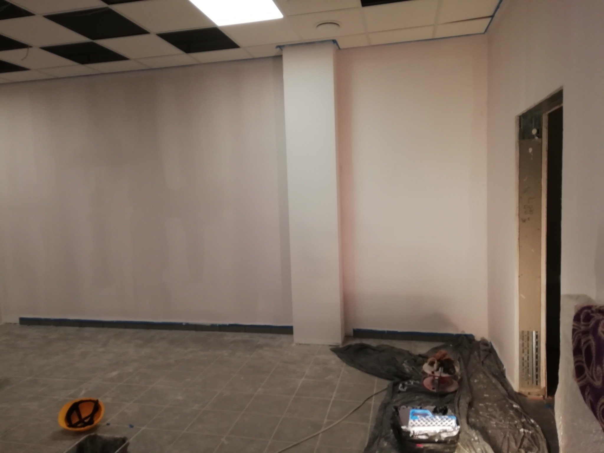 Świeżo pomalowane na biało ściany biura z widoczną taśmą malarską przy suficie, podłodze i kolumnie, z narzędziami malarskimi zabezpieczonymi folią na podłodze oraz żółtym kaskiem w rogu...