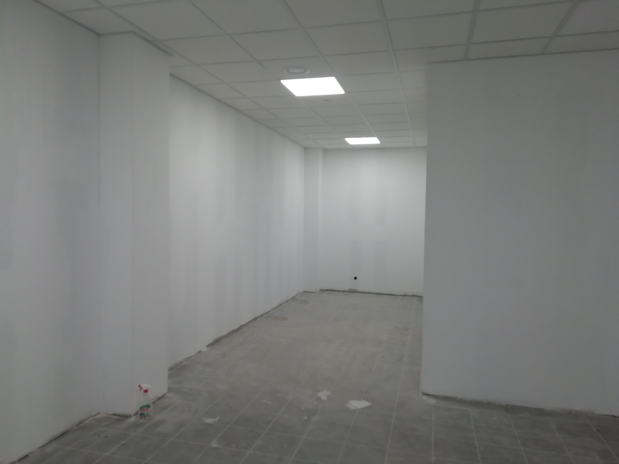 Pomalowane na biało wnętrze biura z kasetonowym sufitem i szarą podłogą w trakcie wykańczania, widoczna butelka ze spryskiwaczem przy ścianie.
