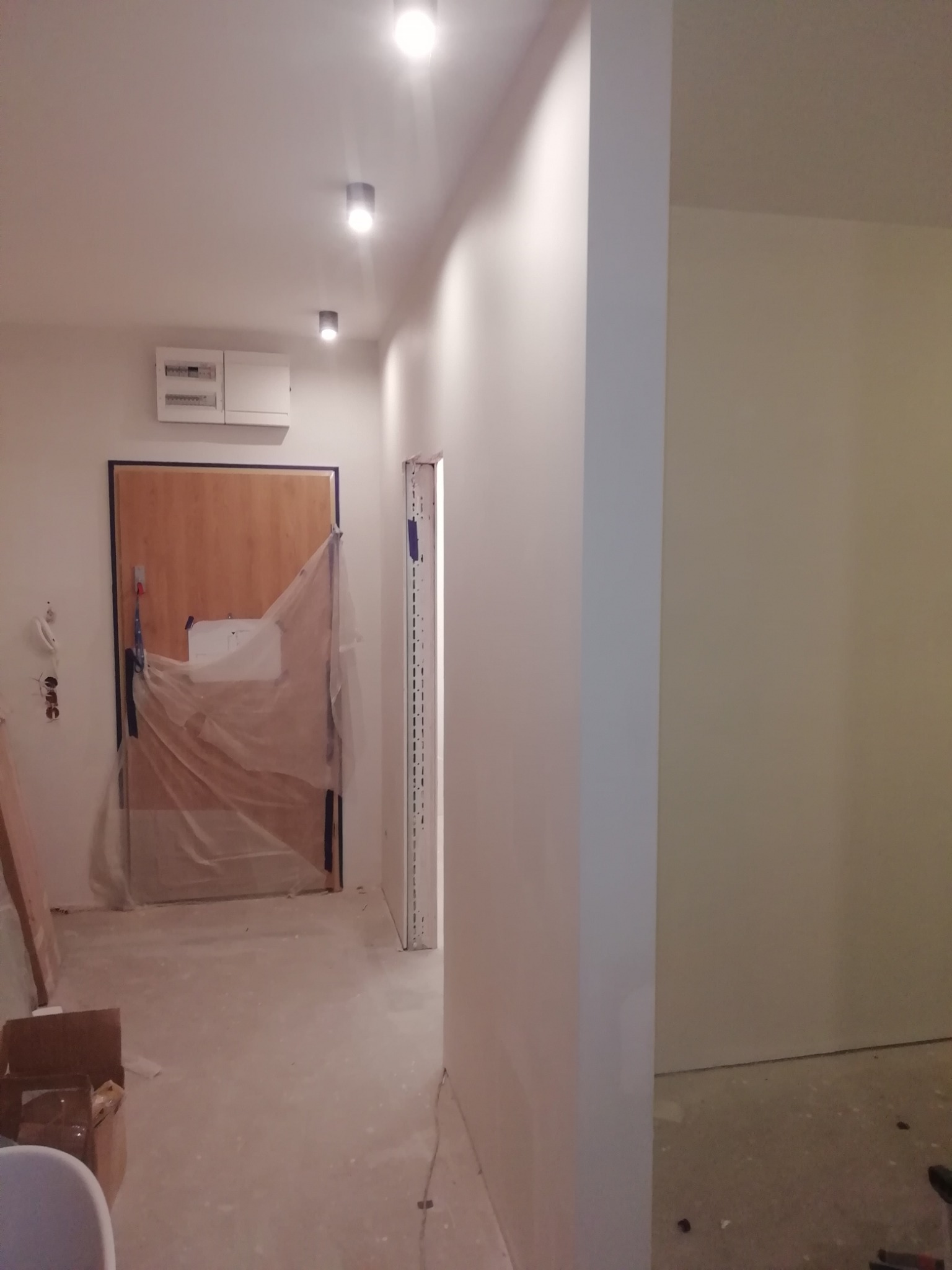 Korytarz w trakcie prac wykończeniowych: zabezpieczone drzwi folią malarską, świeżo pomalowane ściany na biało, widoczne puszki elektryczne i kartony na podłodze.