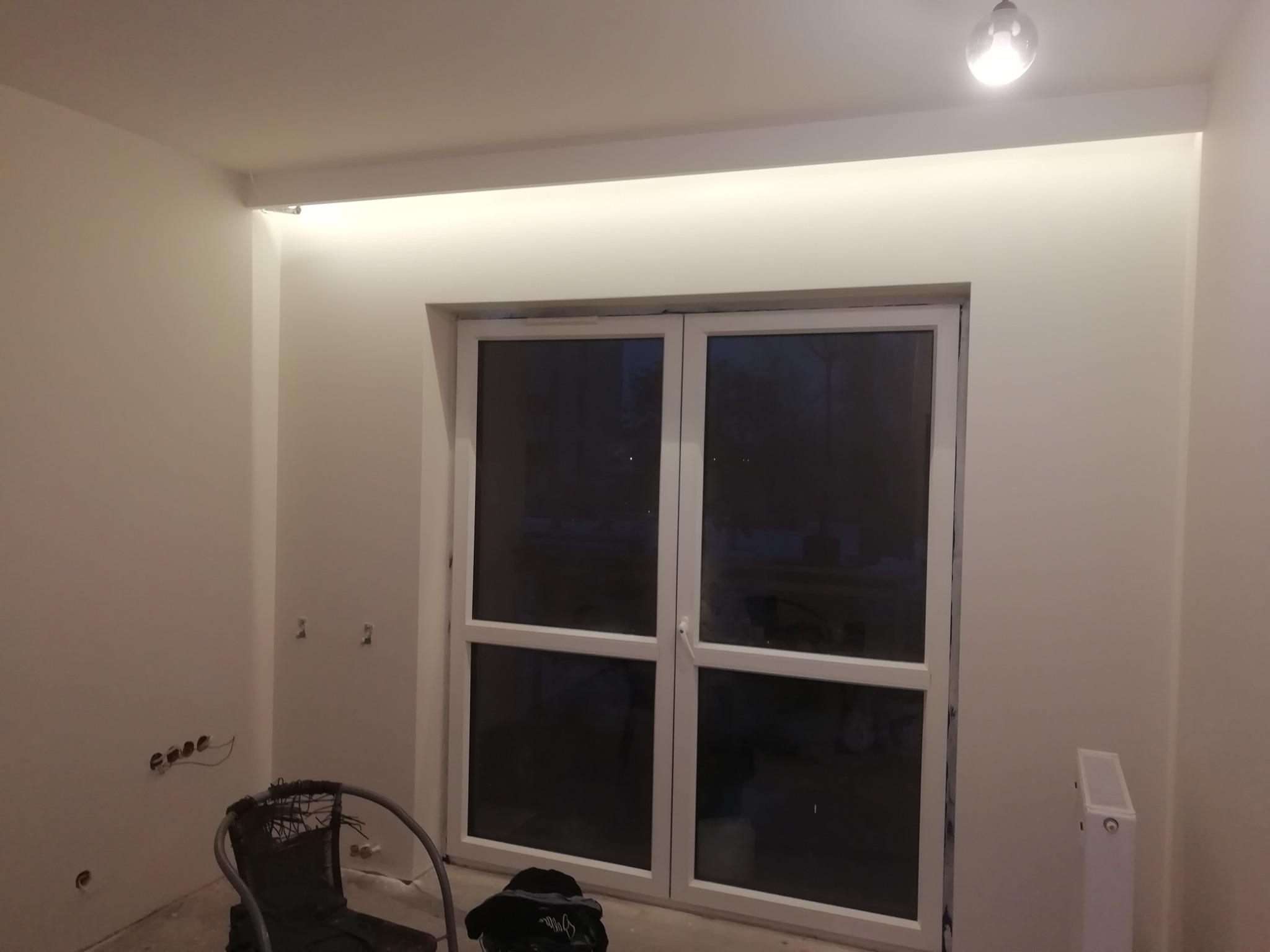 Wnętrze pokoju w trakcie odświeżania, widoczne przygotowane do malowania ściany, okno balkonowe i prowizoryczne oświetlenie sufitowe.