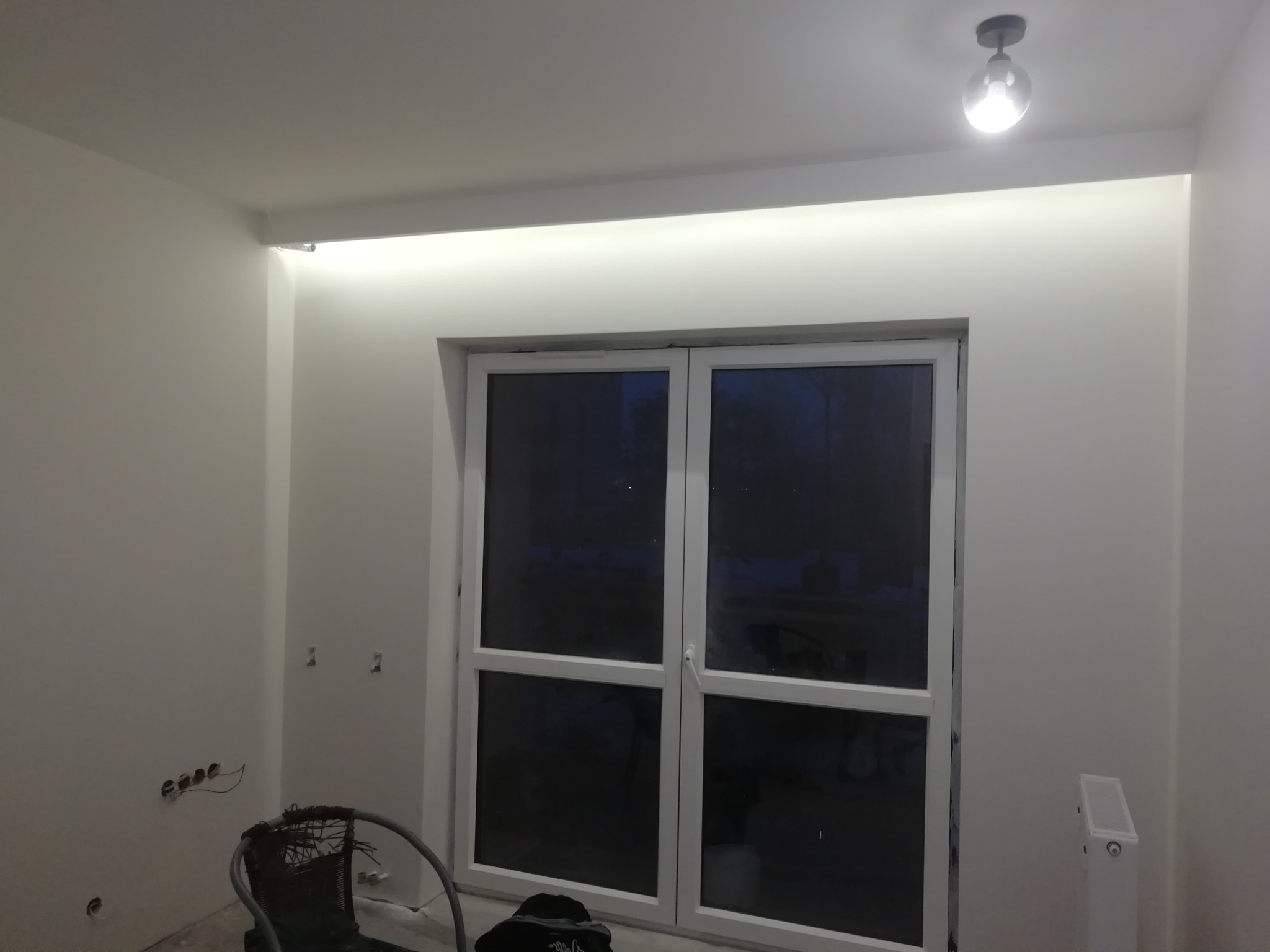 Pokój po malowaniu na biało z podświetleniem LED nad oknem, widoczne gniazdka elektryczne i niedokończone wykończenie.