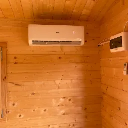Wnętrze drewnianego domku z zamontowaną białą klimatyzacją Haier, wyświetlającą temperaturę 21 stopni Celsjusza, obok skrzynka elektryczna i pilot do klimatyzacji.