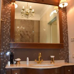 Elegancka łazienka w brązach z drewnianą szafką pod umywalką, złotą armaturą i dużym lustrem z oświetleniem, w którego odbiciu widać kryształowy żyrandol.