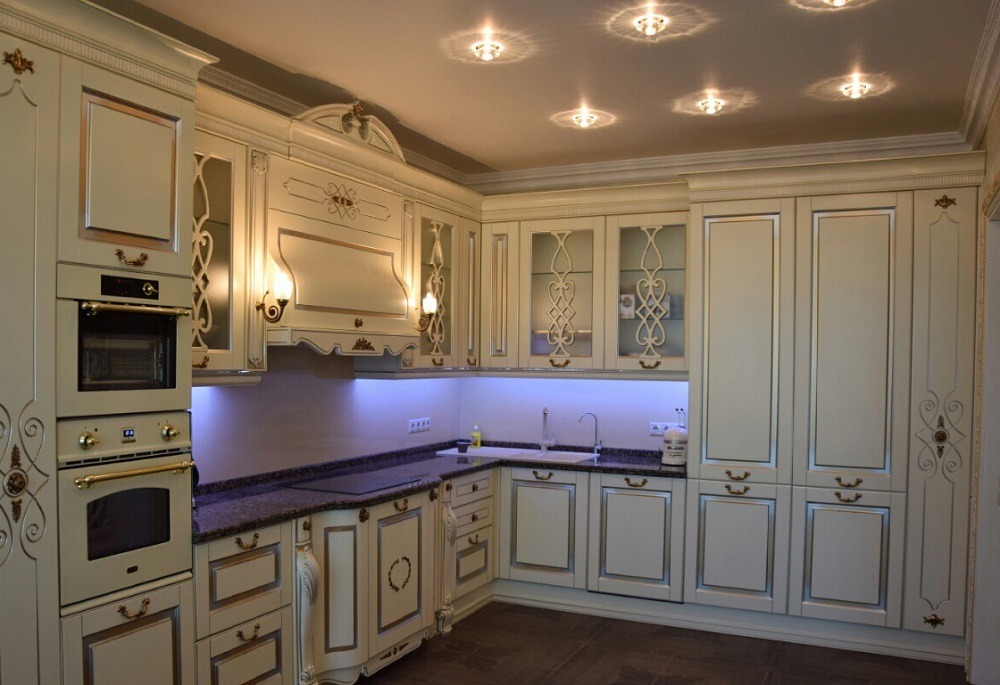 Elegancka kuchnia w stylu klasycznym z kremowymi szafkami zdobionymi złotymi detalami, ciemnym blatem i subtelnym oświetleniem LED pod szafkami wiszącymi.