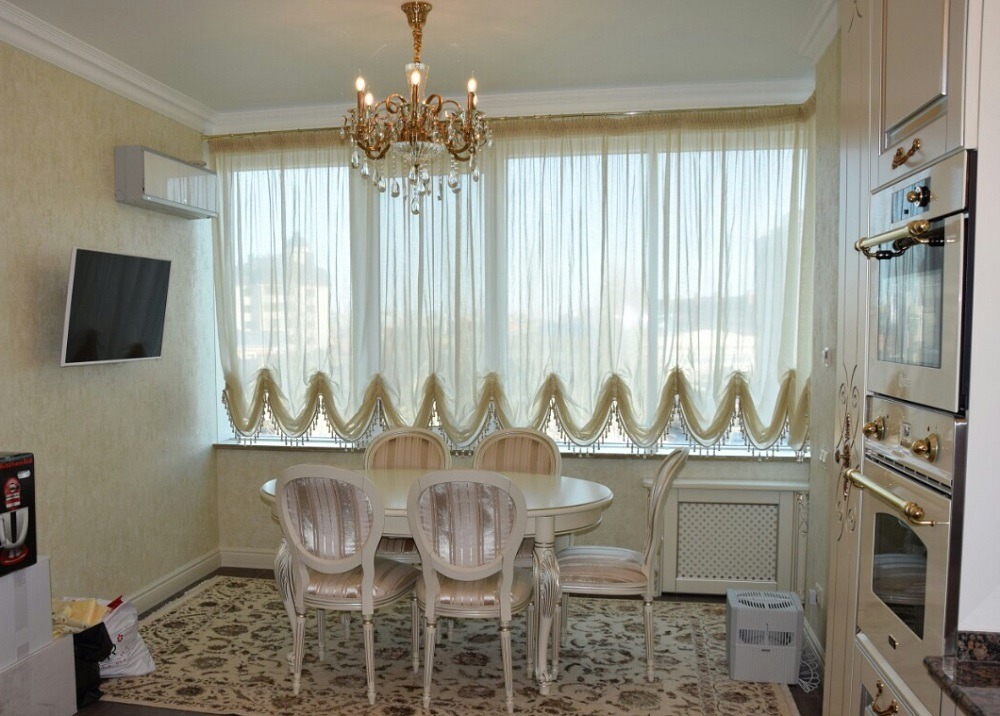 Jasna jadalnia z owalnym stołem i krzesłami w stylu Ludwika XVI, dekoracyjnymi zasłonami z lambrekinami i kryształowym żyrandolem, widok na zabudowę kuchenną i telewizor na ścianie.