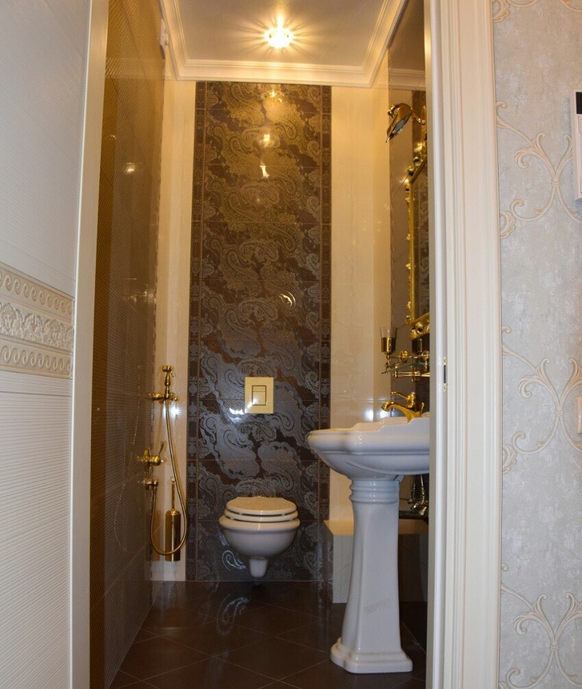 Elegancka łazienka w wąskim pomieszczeniu z ciemną, wzorzystą ścianą z płytek, białą umywalką na postumencie, złotą armaturą i toaletą w stylu retro.