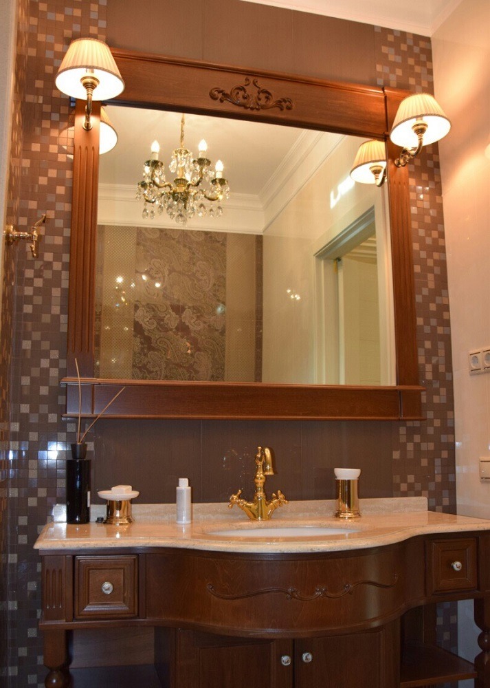 Elegancka łazienka w brązach z drewnianą szafką pod umywalką, złotą armaturą i dużym lustrem z oświetleniem, w którego odbiciu widać kryształowy żyrandol.