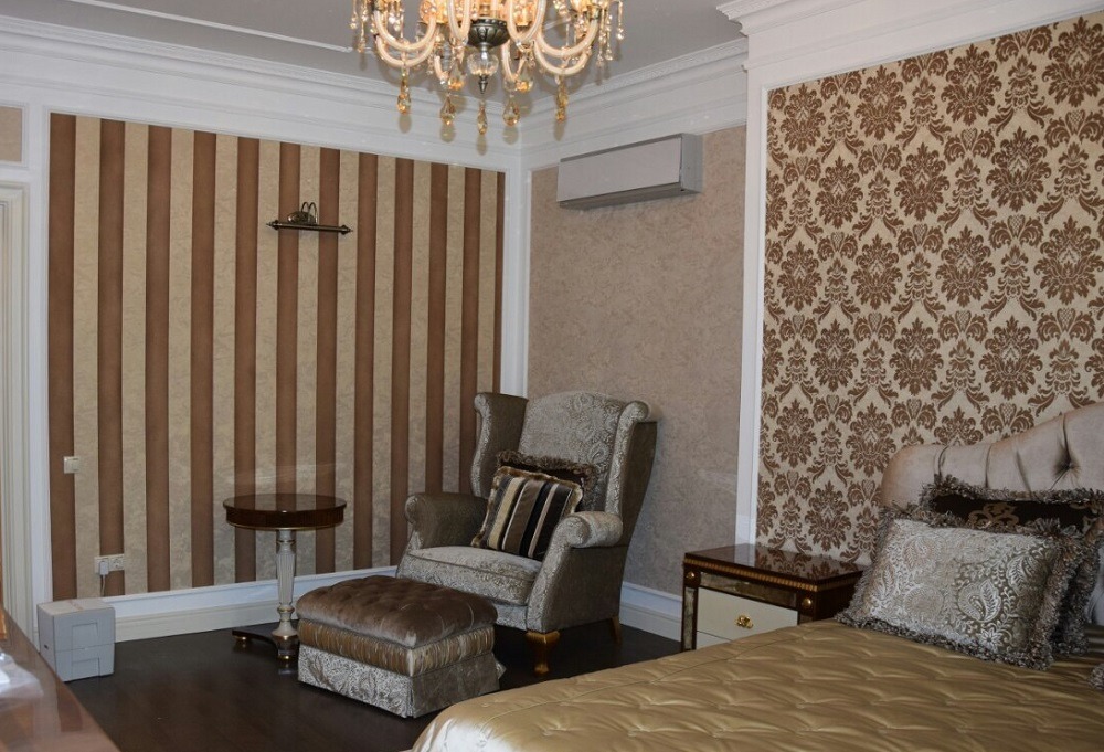 Elegancki pokój z tapetą w paski i wzory, kryształowy żyrandol, fotel z podnóżkiem i łóżko w odcieniach beżu i brązu.