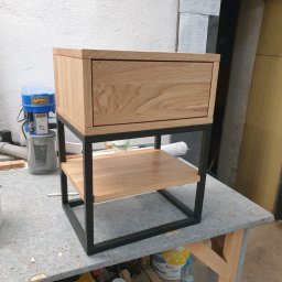 CUSTOM-MET Maciej Franc - Drewniana szafka nocna z szufladą i metalową ramą, stojąca na warsztatowym stole w pomieszczeniu z surowymi ścianami.
