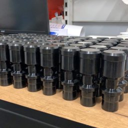 CUSTOM-MET Maciej Franc - Partia czarnych, metalowych, cylindrycznych elementów z kołnierzami, ułożonych rzędami na drewnianym blacie, w tle monitor komputera i dokumenty.