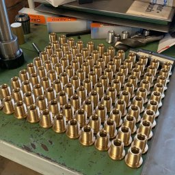 CUSTOM-MET Maciej Franc - Partia precyzyjnie wykonanych, mosiężnych tulejek gwintowanych ułożonych na warsztatowym stole, obok kubek z rysunkiem plażowym i narzędzia.