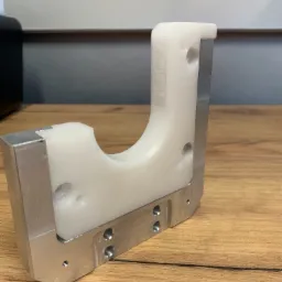 Precyzyjnie wykonany element z aluminium i tworzywa sztucznego, widoczne otwory montażowe i frezowane krawędzie, leżący na drewnianym blacie.