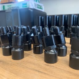 CUSTOM-MET Maciej Franc - Partia czarnych, metalowych złączek hydraulicznych, ustawionych rzędami na drewnianym stole, z widocznym pudełkiem za nimi.