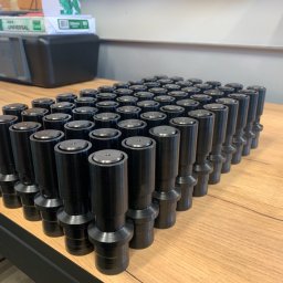 CUSTOM-MET Maciej Franc - Partia czarnych, metalowych elementów cylindrycznych z kołnierzami ułożonych na drewnianym blacie, w tle drukarka i kartony papieru.