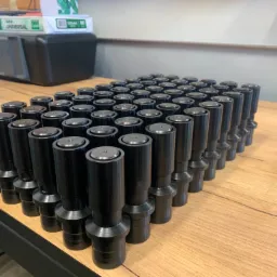 Partia czarnych, metalowych elementów cylindrycznych z kołnierzami ułożonych na drewnianym blacie, w tle drukarka i kartony papieru.