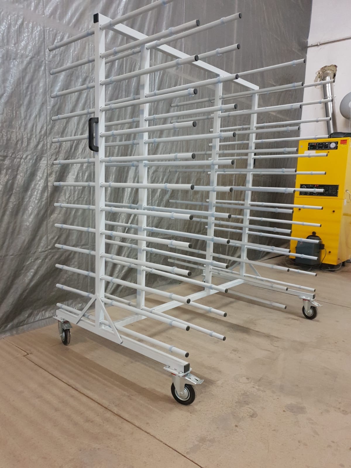 Biały, metalowy regał na kółkach z poziomymi prętami, prawdopodobnie do suszenia elementów po malowaniu, stoi w hali produkcyjnej; w tle widoczna żółta nagrzewnica.