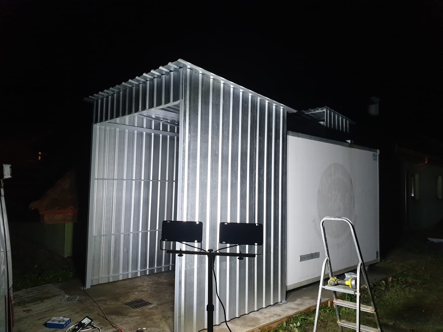 Konstrukcja z blachy falistej, prawdopodobnie wiata lub mała hala, w trakcie budowy nocą, widoczna drabina i narzędzia.