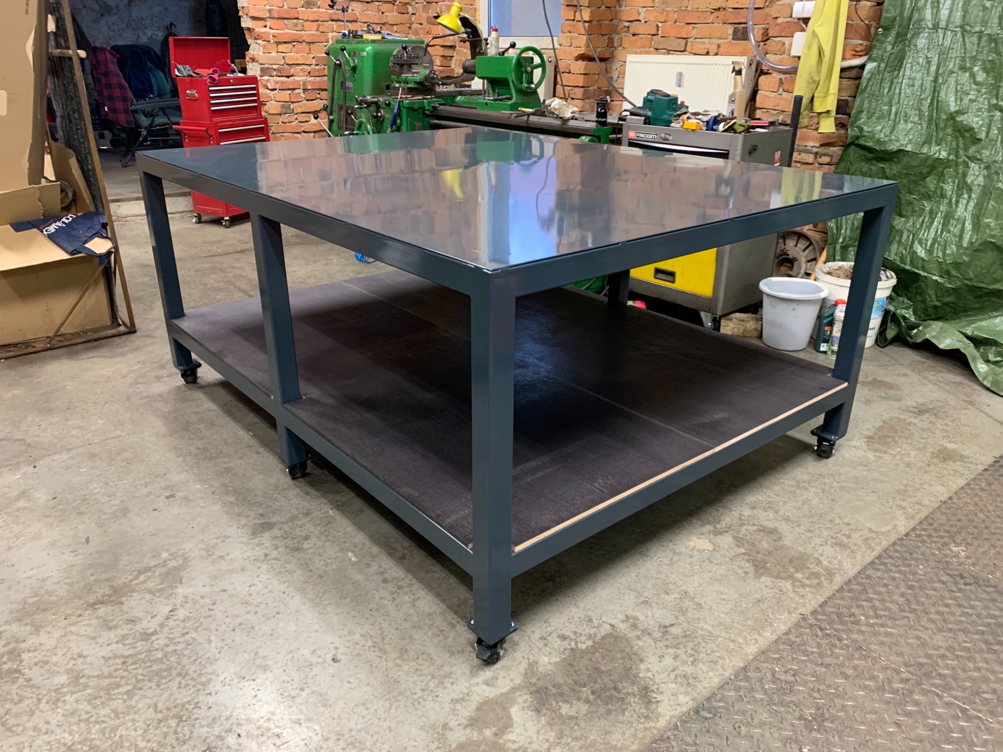 Stół warsztatowy na kółkach z metalową ramą i ciemną, drewnianą półką, w tle ceglana ściana, tokarka i narzędzia.