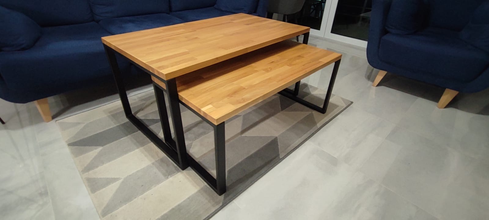 Dwa stoliki kawowe z drewnianymi blatami i czarnymi metalowymi nogami, ustawione jeden pod drugim na dywanie w Twardogórze. W tle widoczna niebieska sofa.