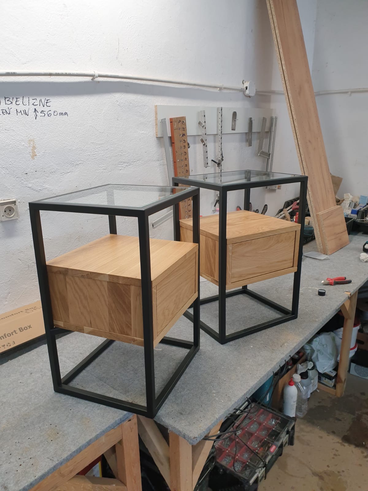 Dwie szafki nocne z drewna dębowego z pojedynczymi szufladami i czarnymi metalowymi ramami, zwieńczone szklanymi blatami, ustawione na warsztatowym stole.