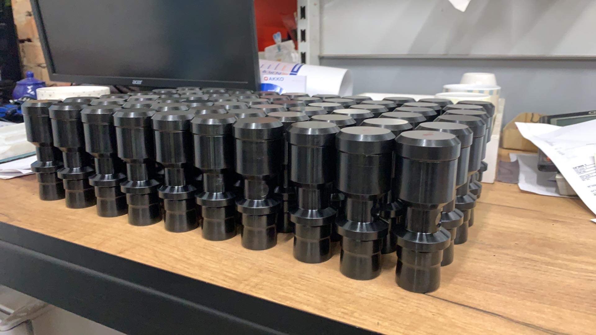 Partia czarnych, metalowych, cylindrycznych elementów z kołnierzami, ułożonych rzędami na drewnianym blacie, w tle monitor komputera i dokumenty.
