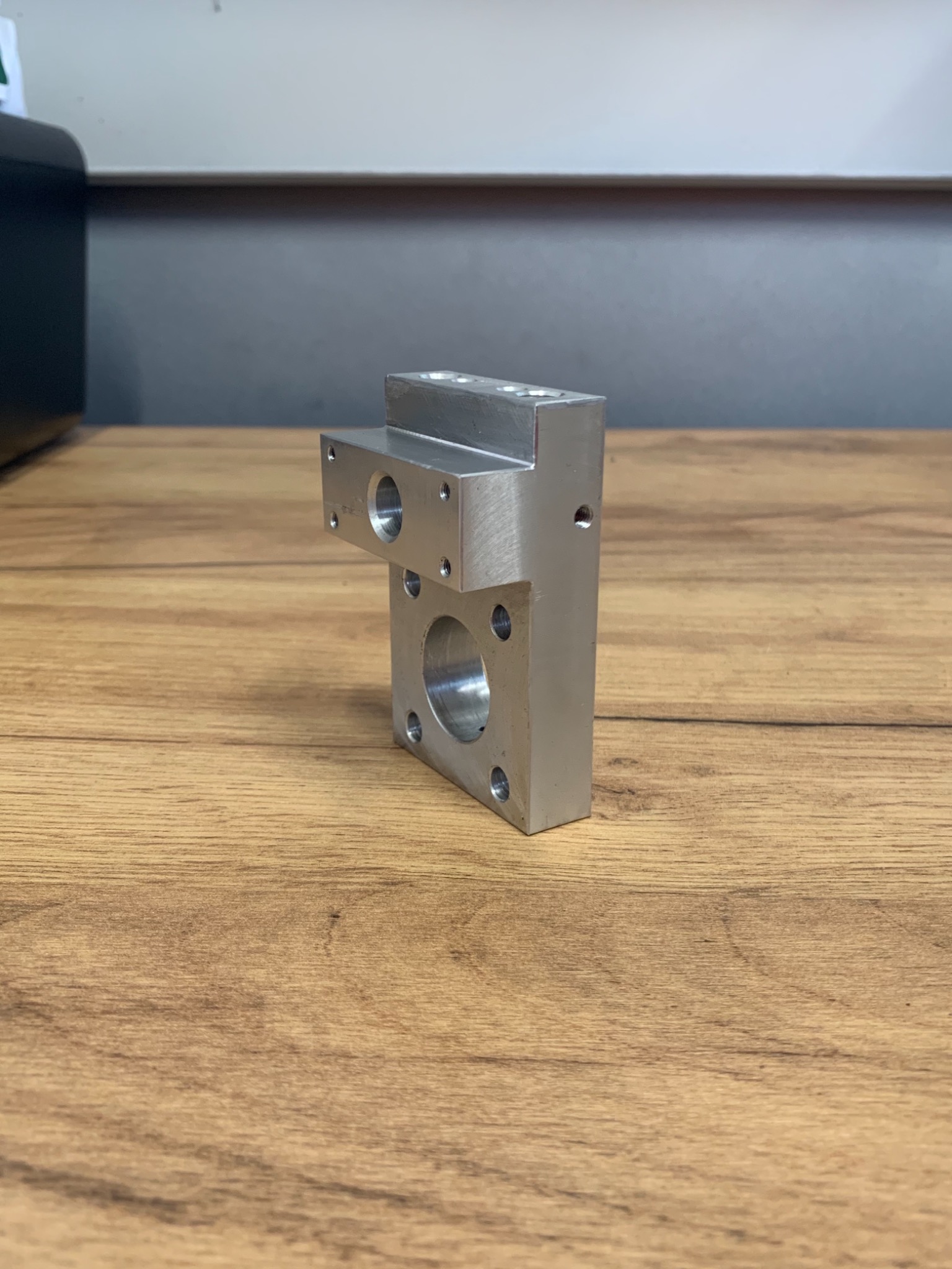 Precyzyjnie obrobiony, srebrny element metalowy z otworami o różnych średnicach, leżący na drewnianej powierzchni.