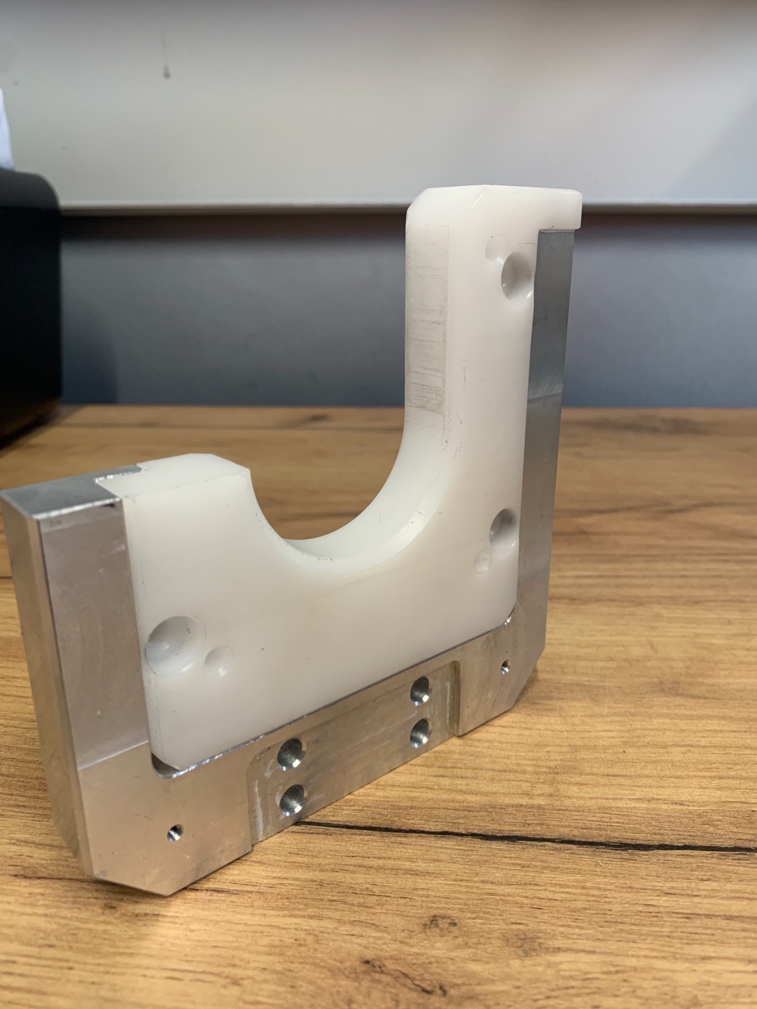 Precyzyjnie wykonany element z aluminium i tworzywa sztucznego, widoczne otwory montażowe i frezowane krawędzie, leżący na drewnianym blacie.