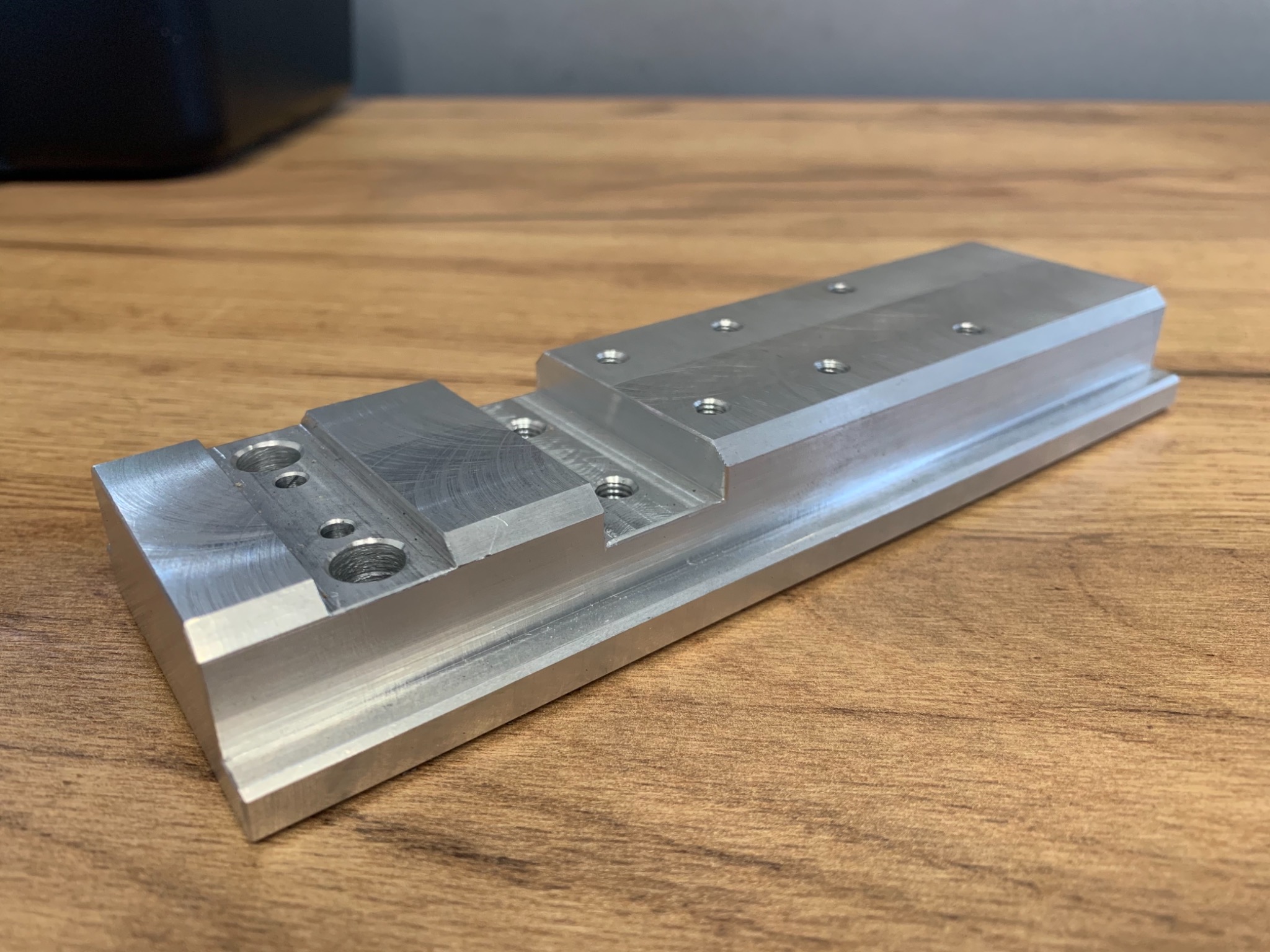 Precyzyjnie obrobiony, aluminiowy element z nawierconymi otworami, leżący na drewnianym blacie. Widoczne ślady obróbki skrawaniem.