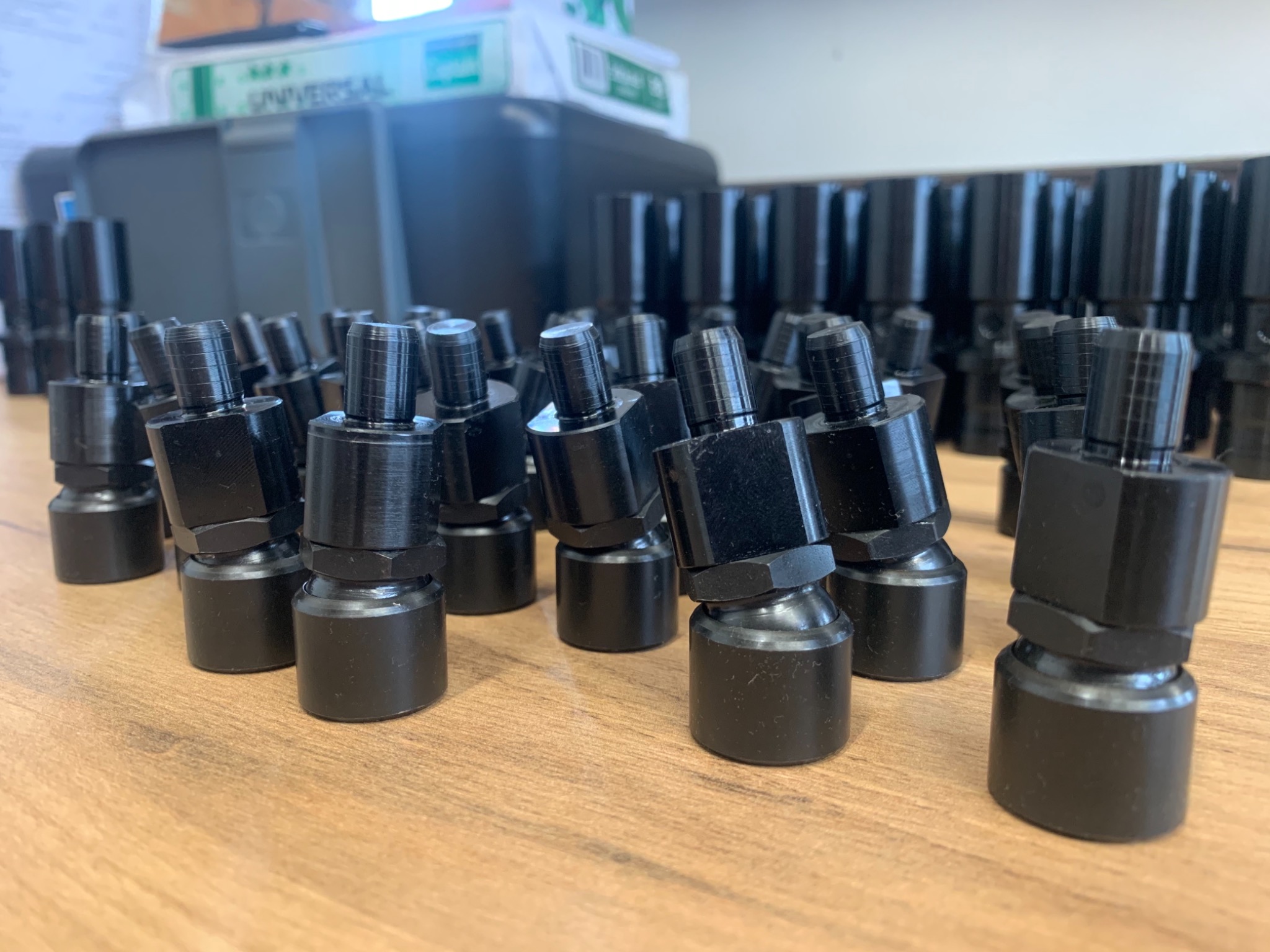 Partia czarnych, metalowych złączek hydraulicznych, ustawionych rzędami na drewnianym stole, z widocznym pudełkiem za nimi.